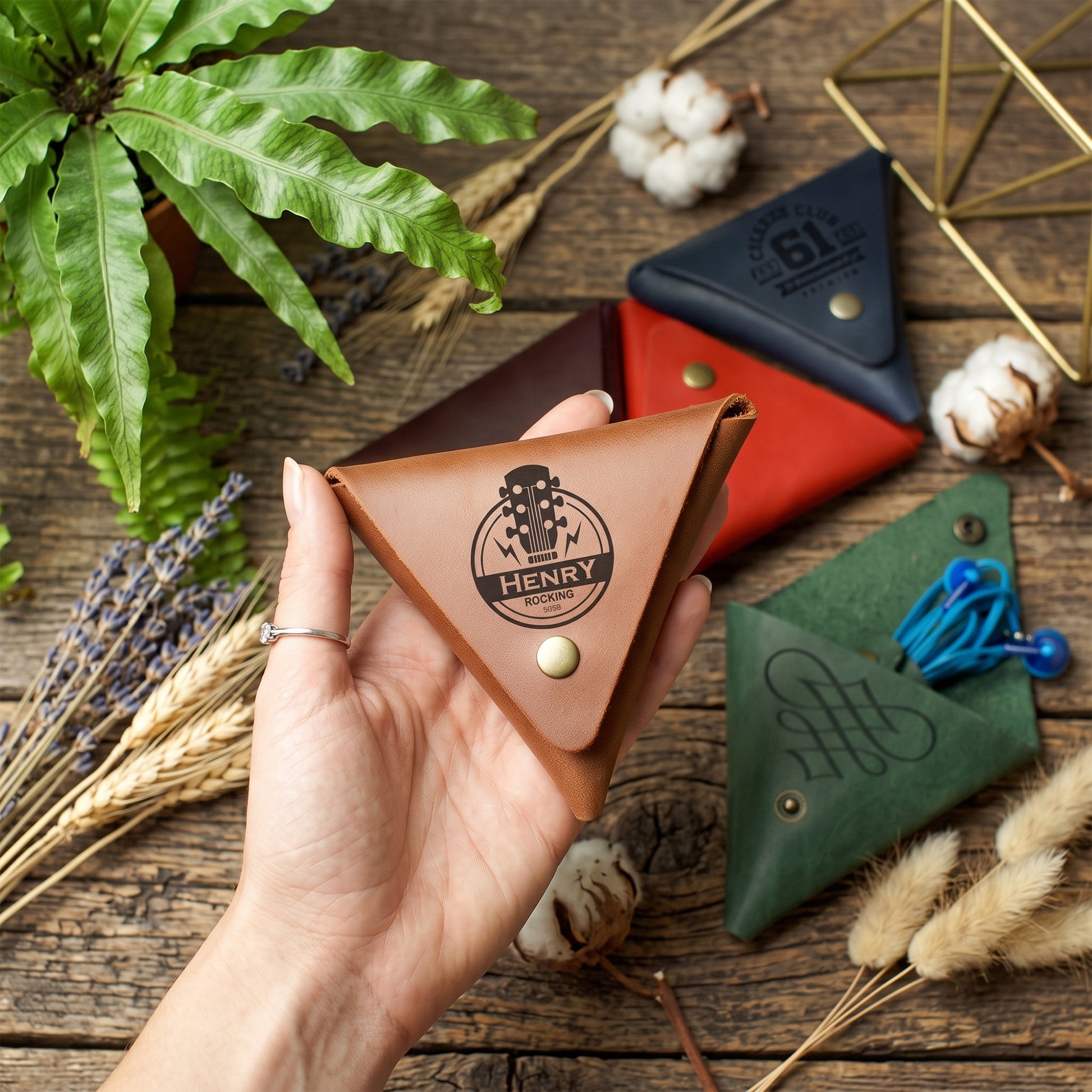A hand holds a triangular brown leather card holder with an engraved logo that reads 'HENRY' and a guitar graphic. Surrounding it are other folded leather card holders in red, navy, and green, along with decorative dried grasses, cotton bolls, a fern, and a gold geometric frame on a rustic wooden surface.