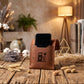 A brown leather phone stand with an embossed fuel pump and lightning bolt design, holding a black smartphone. The stand is placed on a rustic wooden table surrounded by dried flowers, cotton bolls, books, and a warm lamp in the background.