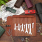 A brown leather wrench roll open on a wooden tabletop, holding several silver wrenches. The scene is styled with a green toy car, a red toy car, wrenches, sketches, dried flowers, and rolled leather sleeves.