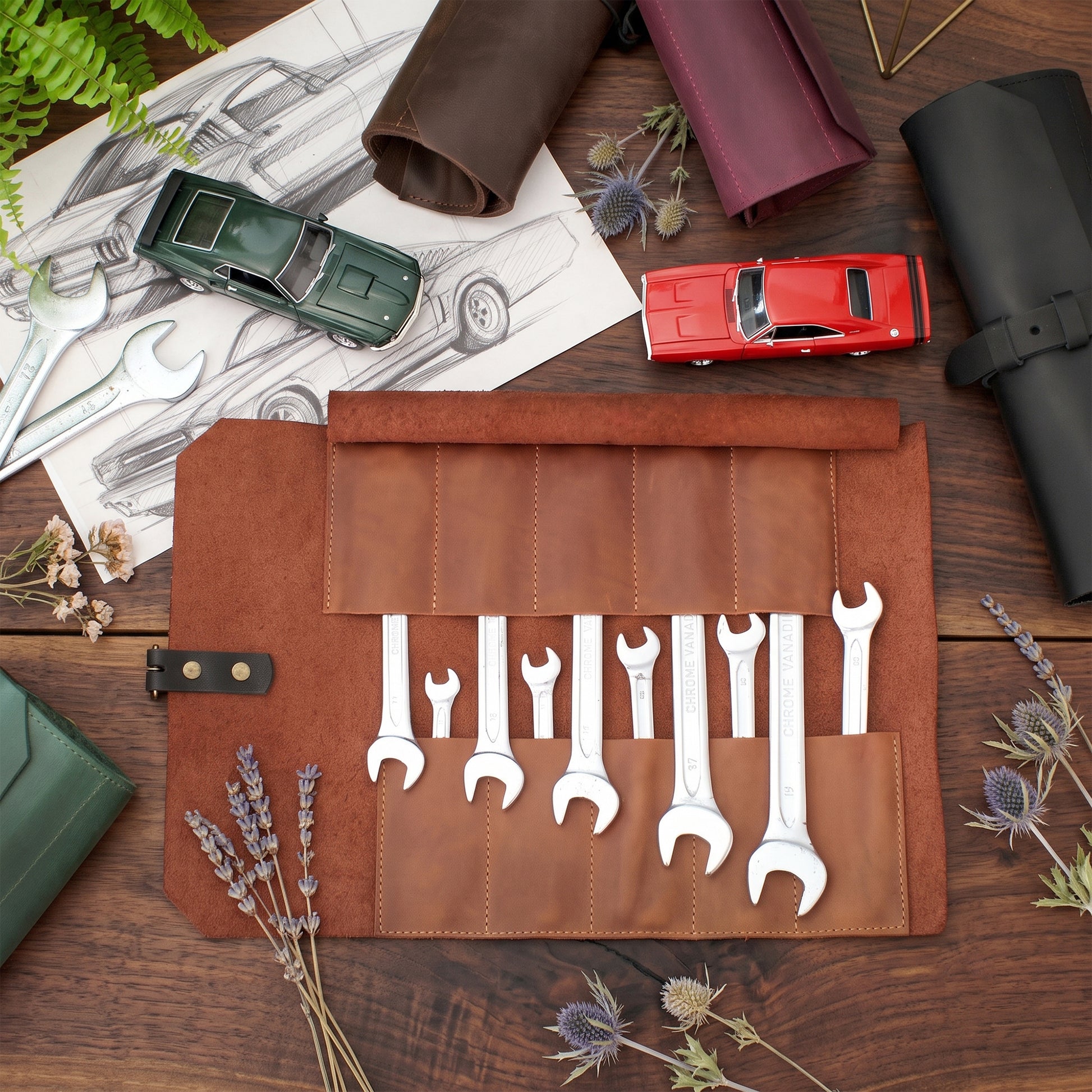 A brown leather wrench roll open on a wooden tabletop, holding several silver wrenches. The scene is styled with a green toy car, a red toy car, wrenches, sketches, dried flowers, and rolled leather sleeves.