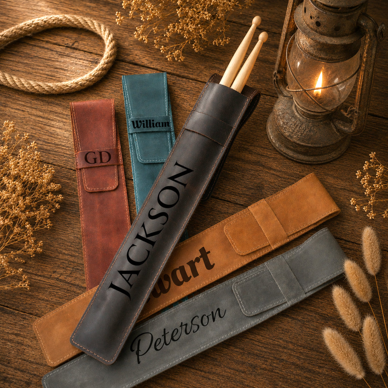 Personalized drumstick pouches and drum sleeves arranged on a rustic wooden surface with dried flowers and a lantern. The set includes a black sleeve with the name JACKSON, a blue sleeve with William, a brown pouch with GD, and a gray sleeve with Peterson, along with matching drumsticks.