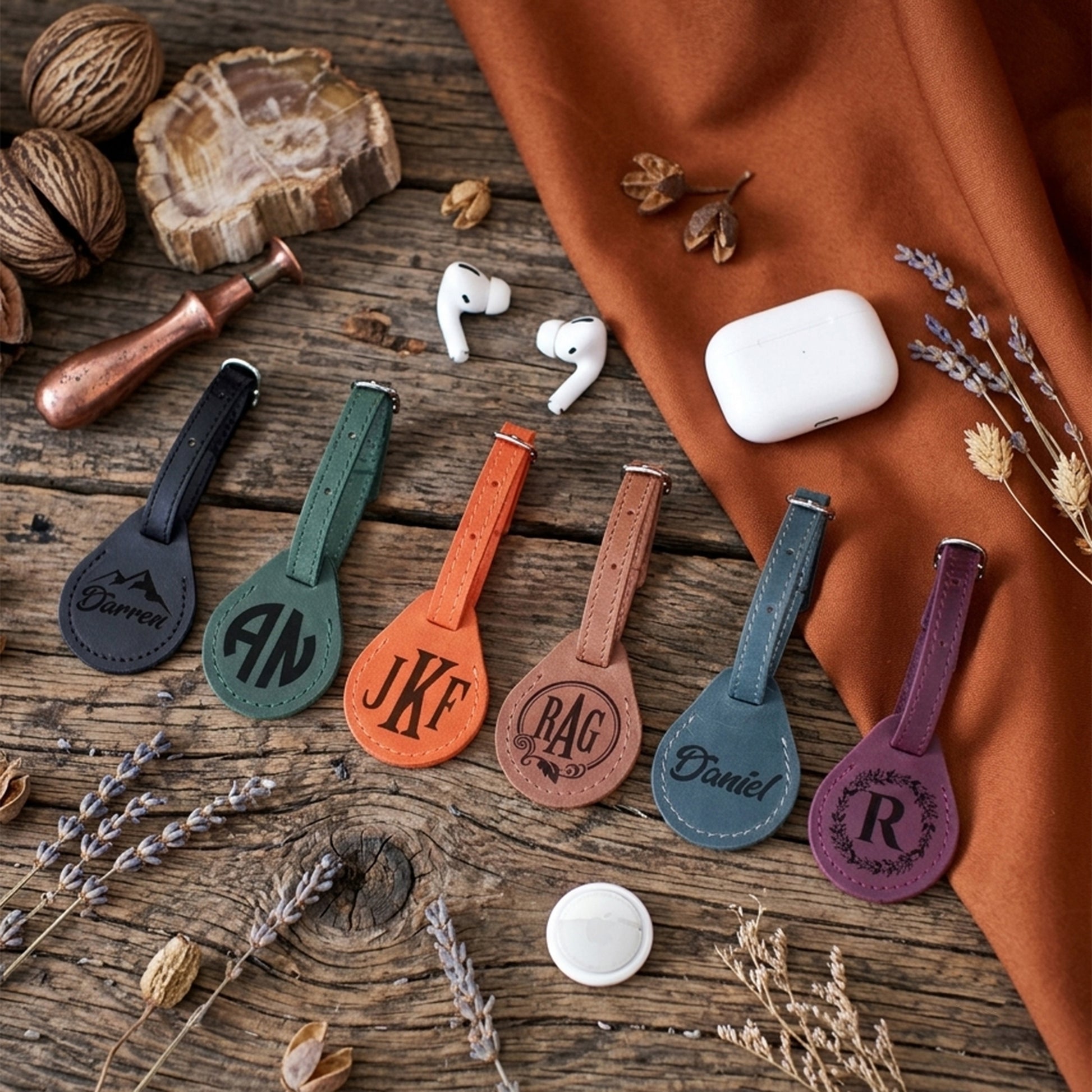 Six personalized leather keychain tags in different colors (black, green, orange, tan, blue, purple) with engraved names or initials, arranged on a rustic wooden table with a white earbud case, white wireless earbuds, a copper funnel, and dried lavender sprigs.