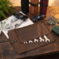A brown leather tool roll laid open on a rustic wooden table, holding several silver wrenches. The scene includes rolled tool cases, toy cars, wrenches, a sketch of a car door, and decorative props, suggesting a styled product presentation.