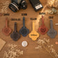 Five personalized leather luggage tags in different colors are arranged on a wooden table with labels above each: Tawny, Chocolate, Denim, Yellow, and Gray. The tags have clover-shaped cutouts, stitched edges, and monogram or name personalization.