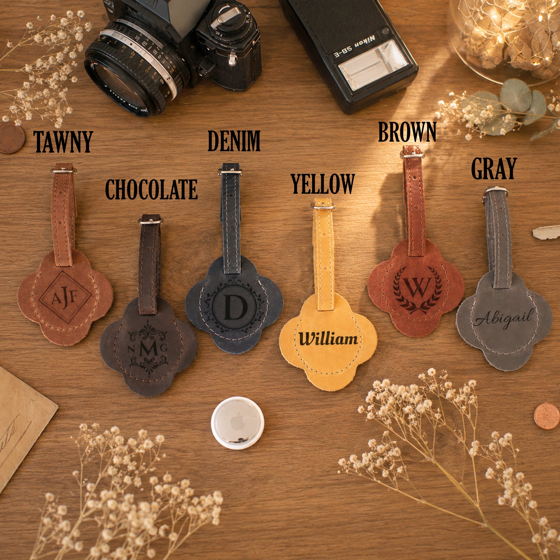Five personalized leather luggage tags in different colors are arranged on a wooden table with labels above each: Tawny, Chocolate, Denim, Yellow, and Gray. The tags have clover-shaped cutouts, stitched edges, and monogram or name personalization.