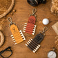Three personalized leather golf keychains styled on a wooden tabletop with props including a golf ball, camera, glasses, pocket watch, rope, and dried flowers. The keychains are tan, reddish-brown, and dark brown, each with a metal clip and a row of white-tipped simulated golf clubs.