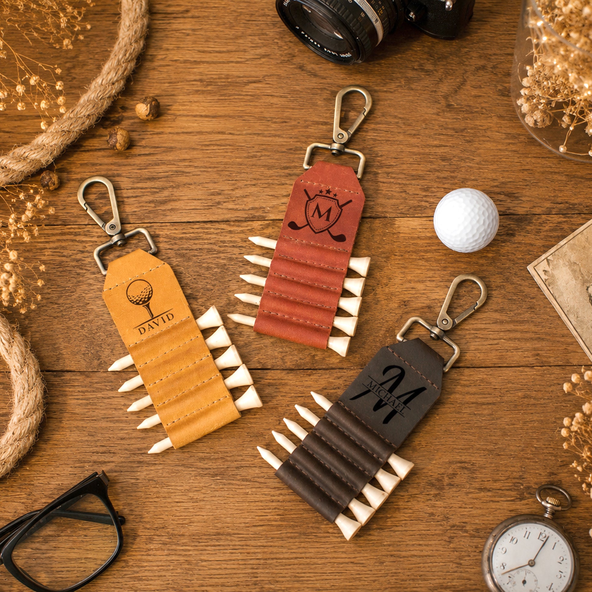 Three personalized leather golf keychains styled on a wooden tabletop with props including a golf ball, camera, glasses, pocket watch, rope, and dried flowers. The keychains are tan, reddish-brown, and dark brown, each with a metal clip and a row of white-tipped simulated golf clubs.