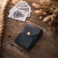 A small dark leather card holder or wallet with a gold snap button, placed on a rustic wooden surface beside playing cards and dried flowers.