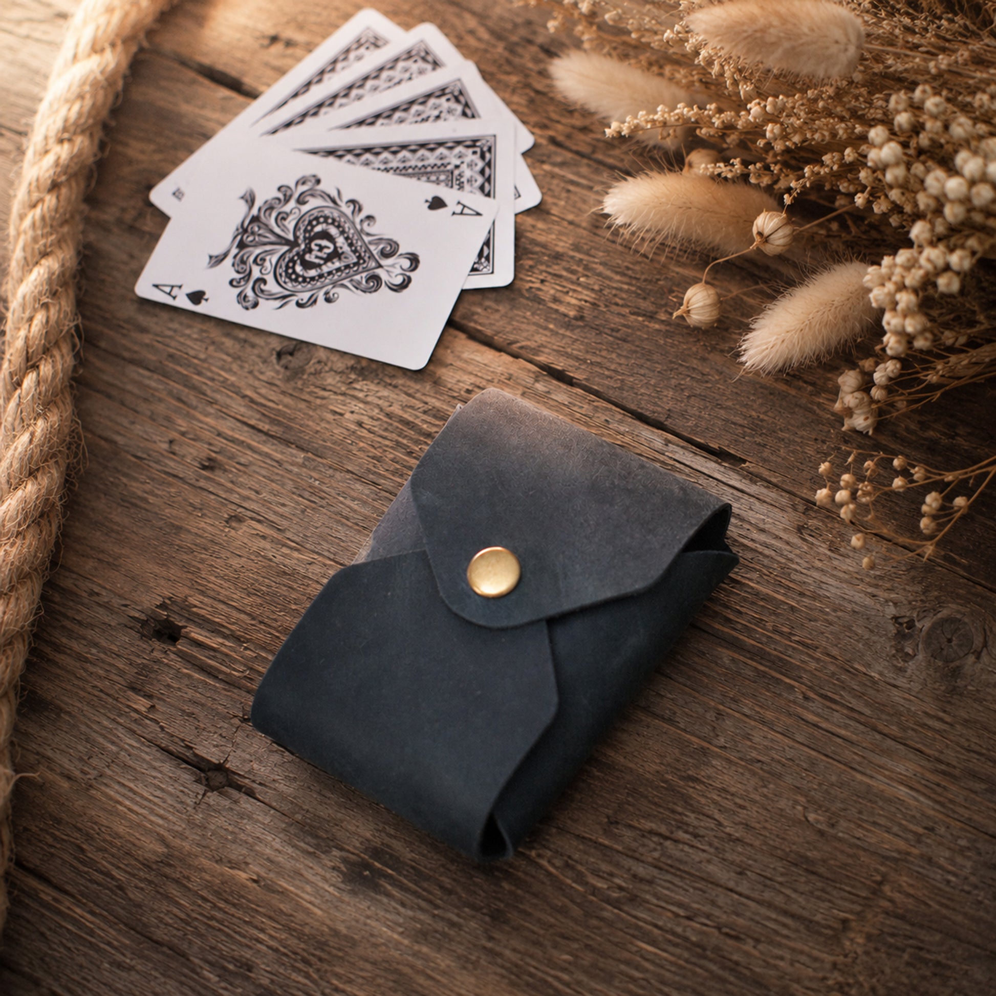 A small dark leather card holder or wallet with a gold snap button, placed on a rustic wooden surface beside playing cards and dried flowers.