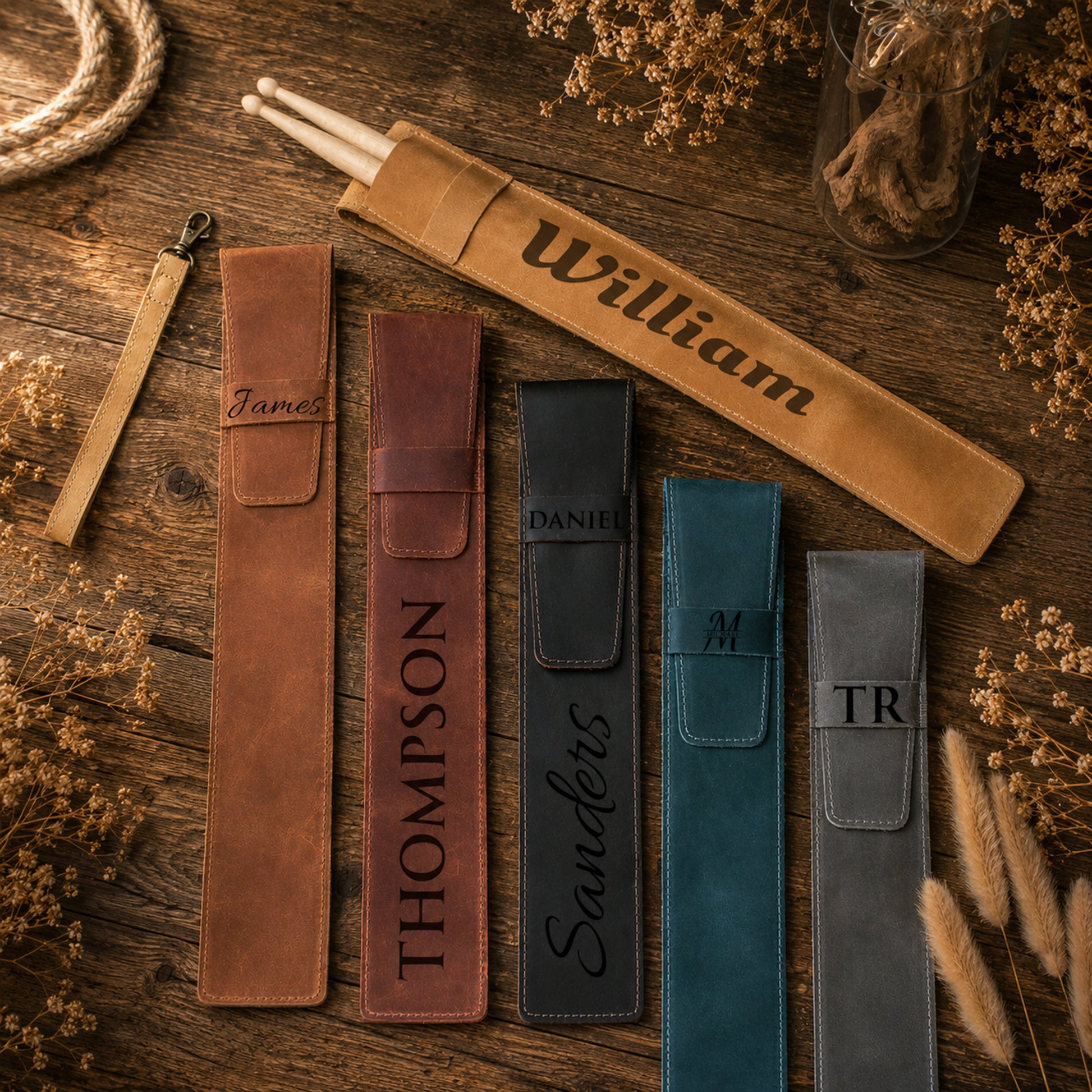 Five personalized leather pencil cases in brown, reddish-brown, black, teal, and gray, laid out on a rustic wooden surface with dried flowers and rope props. The cases are engraved with names including William, James, Thompson, Sanders, and TR.