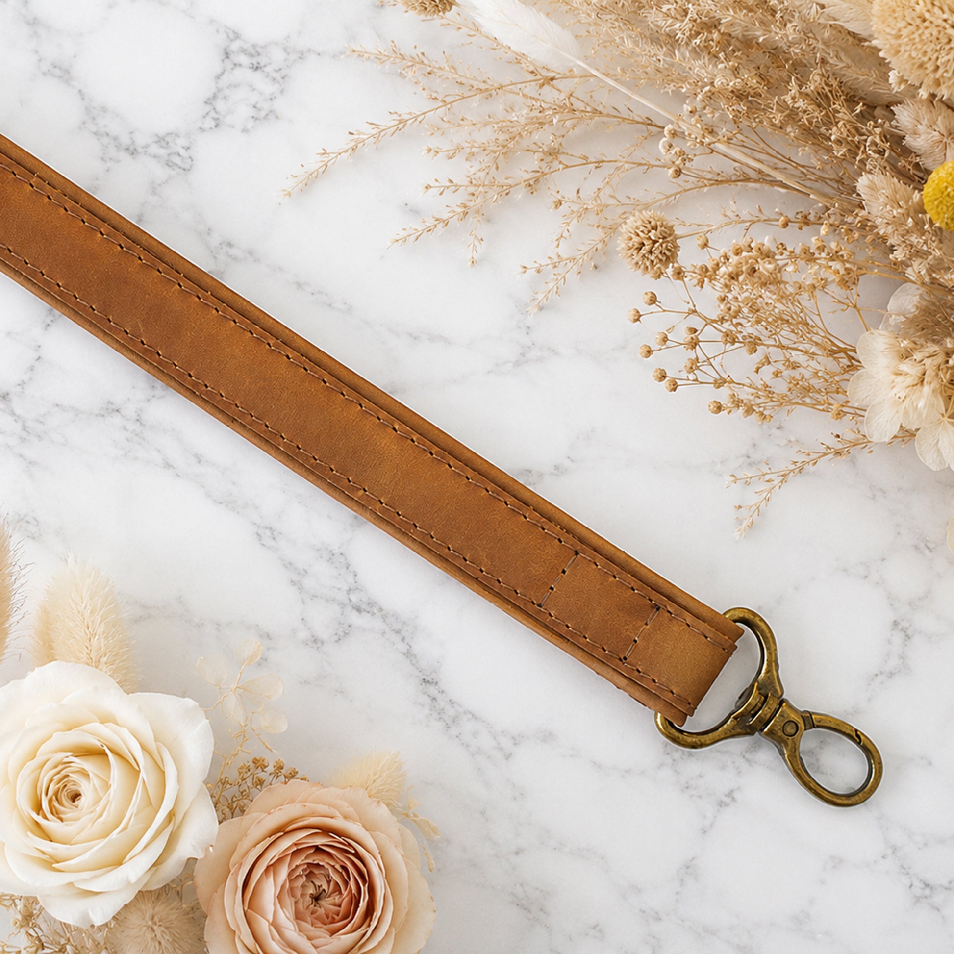 Brown leather strap with brass hook on a marble surface with flowers and dried plants.