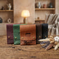 Four leather card sleeves or wallet-style cases in burgundy, green, tan, and black, arranged on a rustic wooden table with dried flowers and a softly blurred indoor room in the background. The tan case in front has the brand name 'Muriel' printed on it.