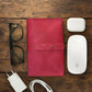 Pink leather cable organizer, glasses, white Apple products on a wooden surface