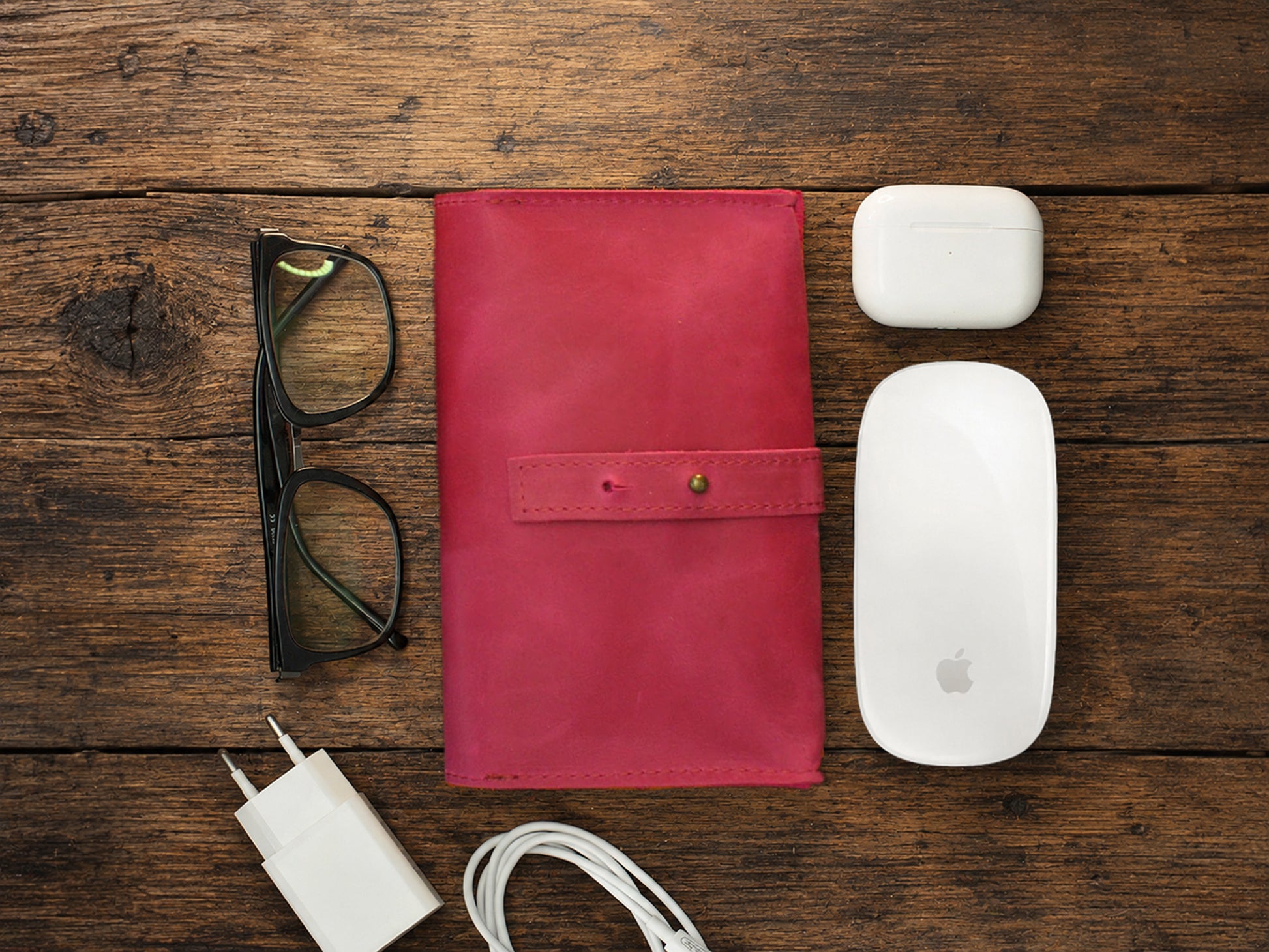 Pink leather cable organizer, glasses, white Apple products on a wooden surface
