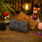 A personalized dark gray suede crocheted clutch case and a brown leather crochet envelope-style pouch, both engraved with names, displayed on a rustic wooden table surrounded by crochet props, yarn balls, a crocheted teddy bear, and a vintage lantern.