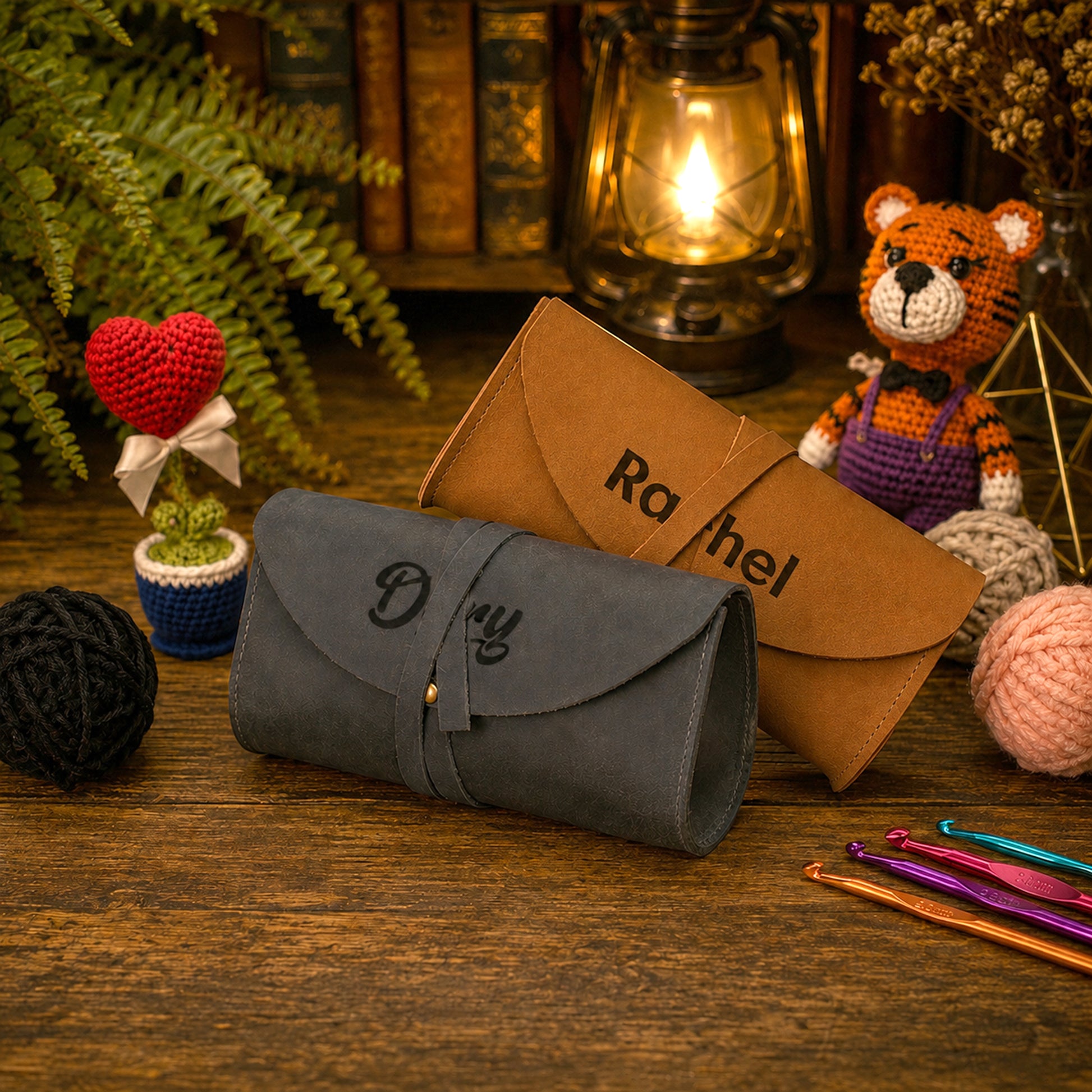 A personalized dark gray suede crocheted clutch case and a brown leather crochet envelope-style pouch, both engraved with names, displayed on a rustic wooden table surrounded by crochet props, yarn balls, a crocheted teddy bear, and a vintage lantern.