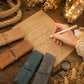 A styled flat-lay of personalized leather ring binders on a rustic wooden table, with a hand holding a white pen writing on a parchment paper. The binders come in tan, slate blue, and gray leather, each customized with a different name (Bessie, William, and Anabel) in black script.