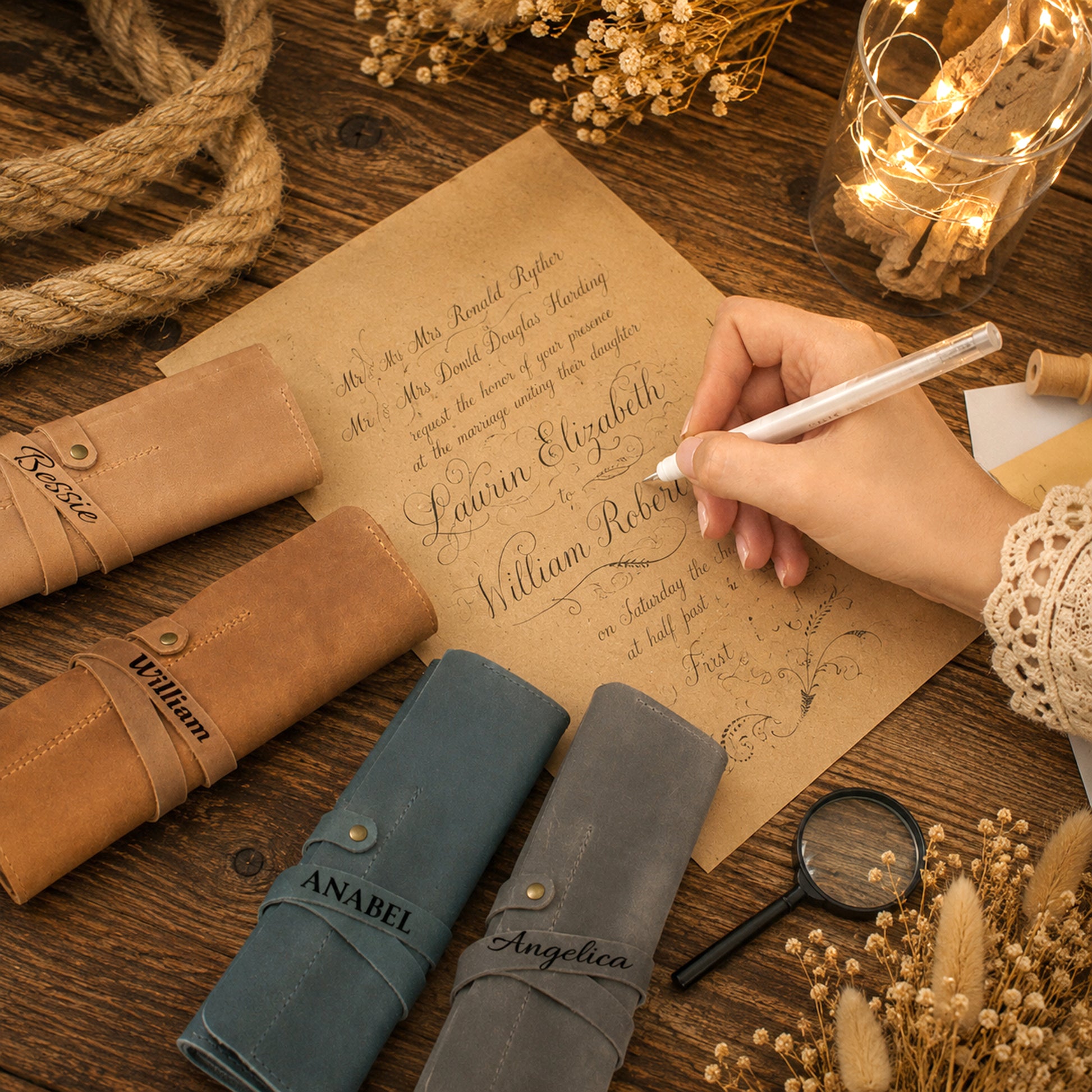 A styled flat-lay of personalized leather ring binders on a rustic wooden table, with a hand holding a white pen writing on a parchment paper. The binders come in tan, slate blue, and gray leather, each customized with a different name (Bessie, William, and Anabel) in black script.