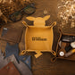 A personalized tan leather dog toy tray with the name 'William' printed on the front, styled in a flat-lay arrangement on a wooden table with sunglasses, a wallet, keys, and leather samples.