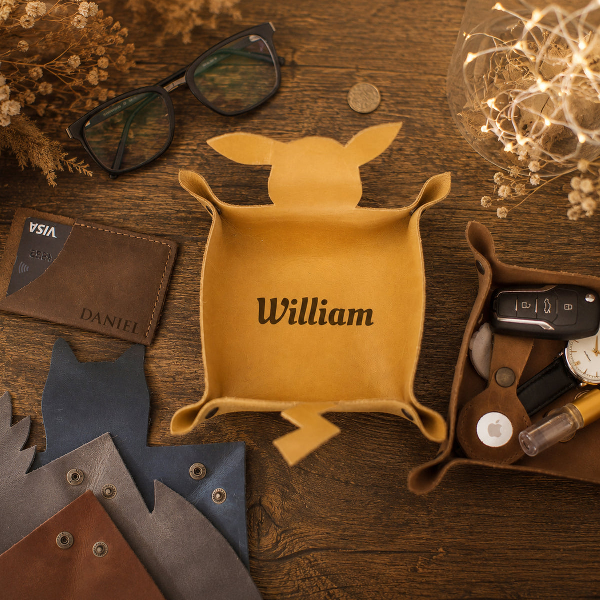 A personalized tan leather dog toy tray with the name 'William' printed on the front, styled in a flat-lay arrangement on a wooden table with sunglasses, a wallet, keys, and leather samples.