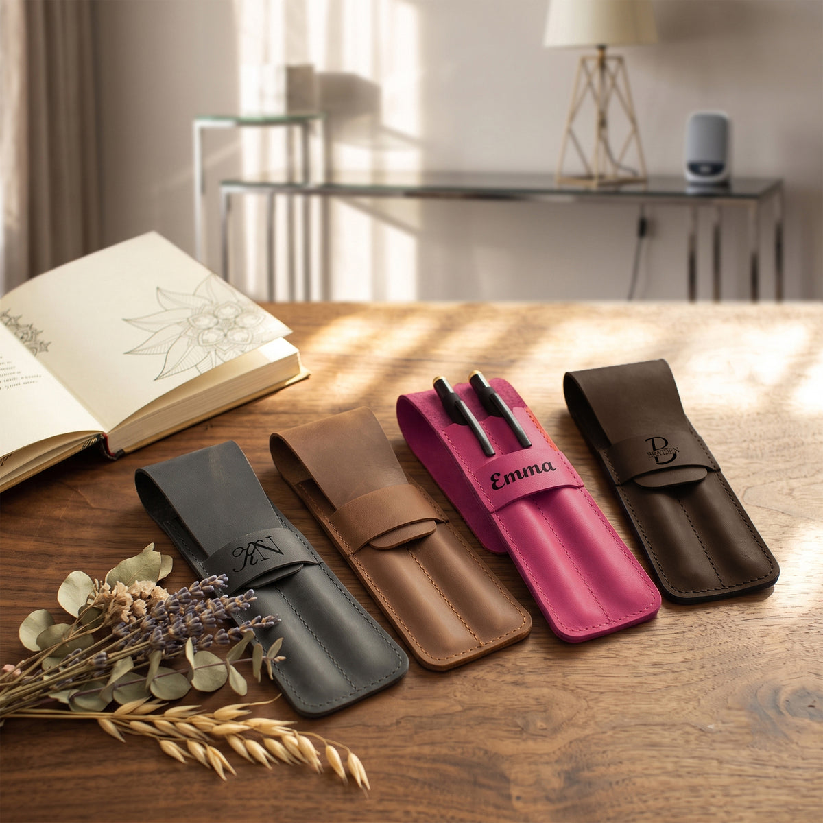 Four personalized leather pen cases in black, brown, tan, and pink, arranged on a wooden table with dried wheat and eucalyptus sprigs. The pink case is open, displaying two black pens, and the cases feature monogrammed straps with initials 'M', 'E', 'B', and 'K'.