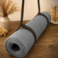 A rolled gray textured yoga mat secured with black straps, resting on a wooden table with dried flowers and a vase in the background.