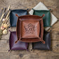A set of square leather storage bins or trays with rounded corners, arranged on a rustic wooden surface. A central brown bin is open and features the words 'Home Sweet Home' printed on the inside.