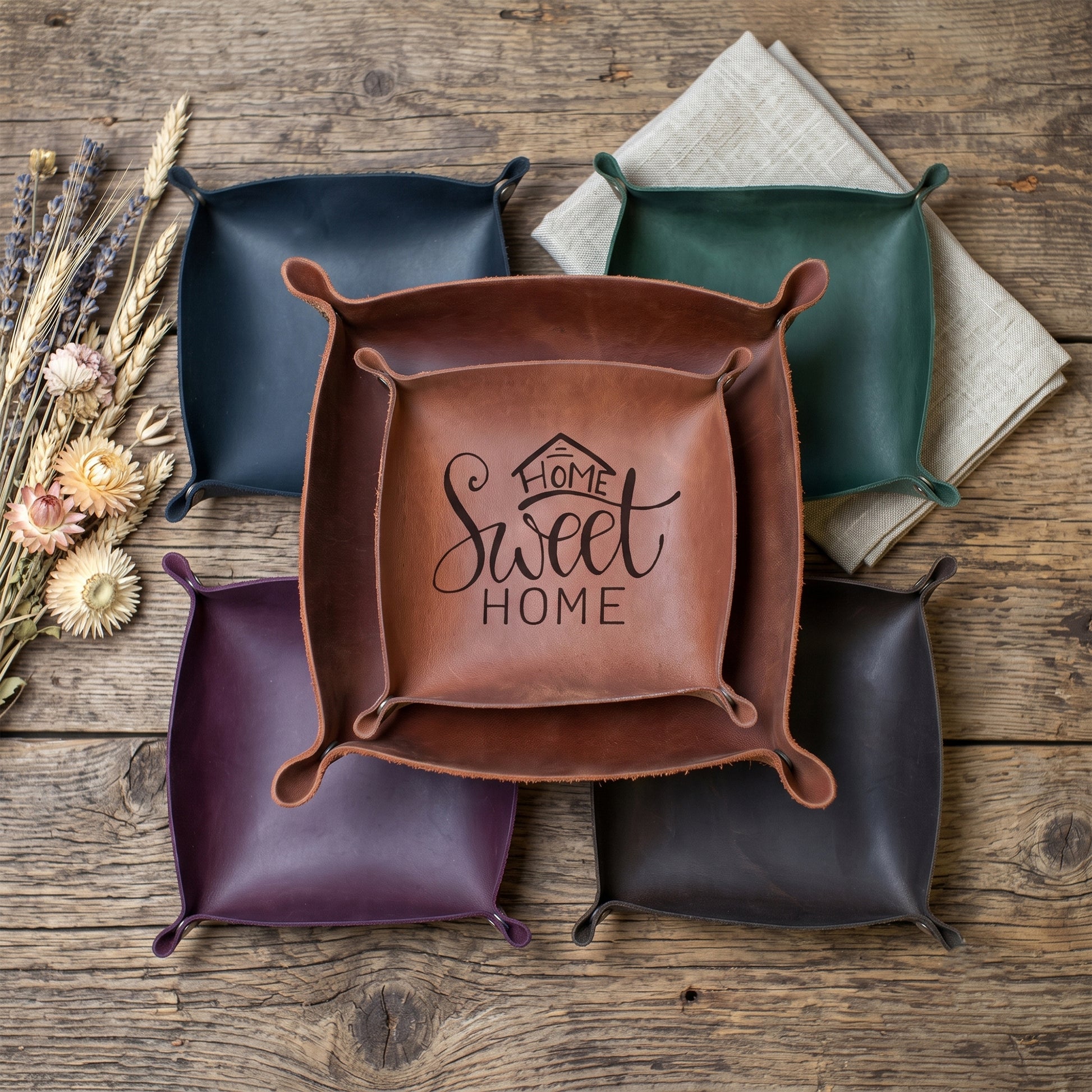 A set of square leather storage bins or trays with rounded corners, arranged on a rustic wooden surface. A central brown bin is open and features the words 'Home Sweet Home' printed on the inside.
