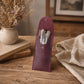 A burgundy leather makeup brush pouch standing upright on a wooden table, holding several makeup brushes. The pouch is styled in a warm, rustic setting with dried flowers in a speckled vase, stacked books, and a beige linen cloth.