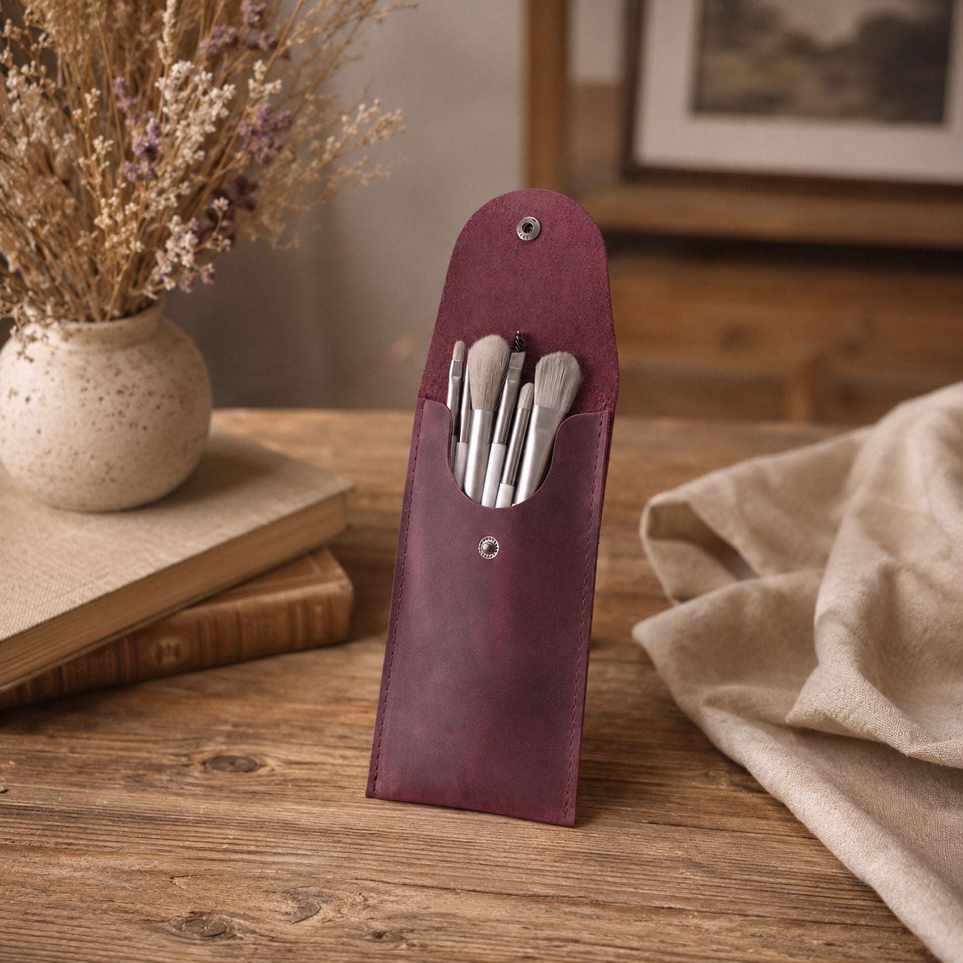 A burgundy leather makeup brush pouch standing upright on a wooden table, holding several makeup brushes. The pouch is styled in a warm, rustic setting with dried flowers in a speckled vase, stacked books, and a beige linen cloth.