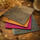 Four suede-like card sleeves or wallet-like accessories stacked on a rustic wooden surface, with dried flowers and a glass jar nearby. The visible colors are taupe-gray, tan, magenta, and slate blue, each with metal snap buttons.