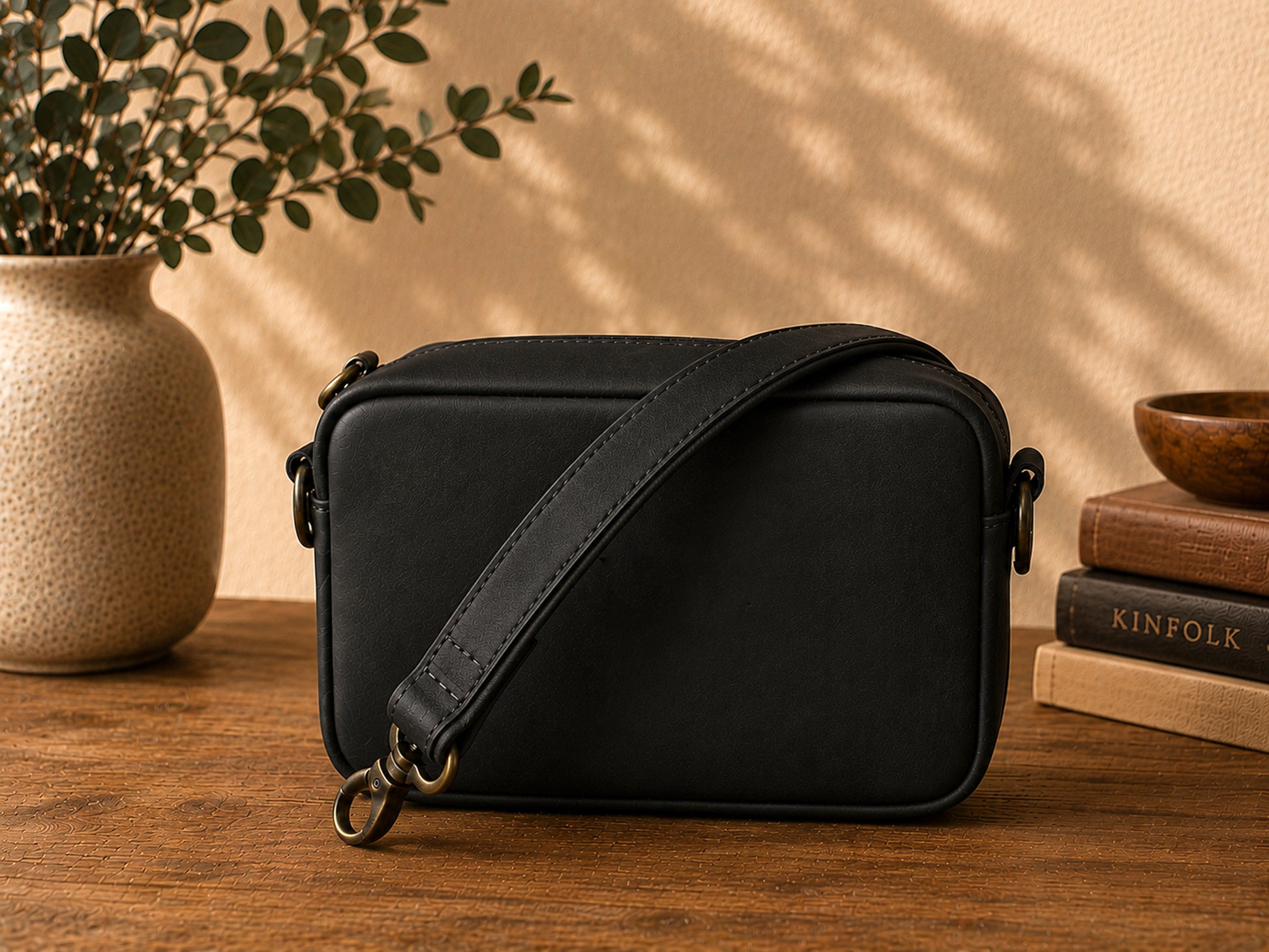 Black leather crossbody bag on a wooden surface with books and a plant in the background