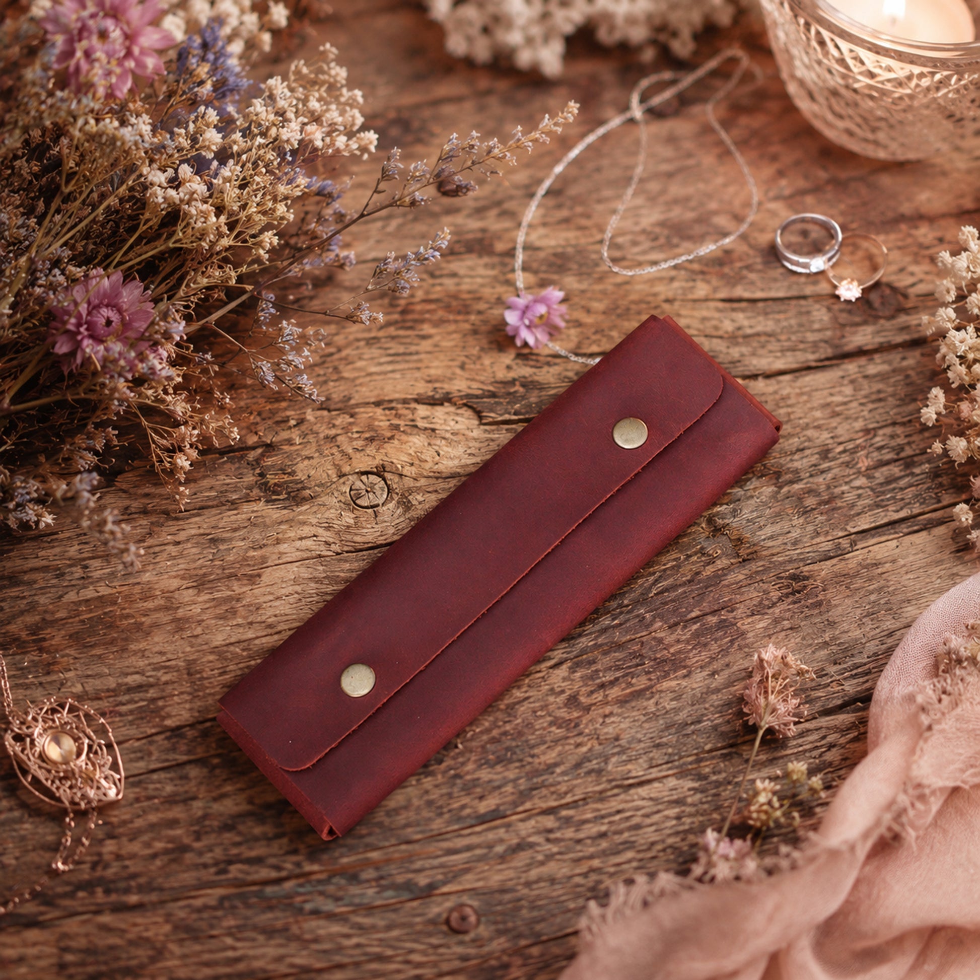 A dark reddish-brown leather ring roll or wallet with two gold-tone snap buttons, styled on a rustic wooden surface with dried flowers, rings, and a candle.