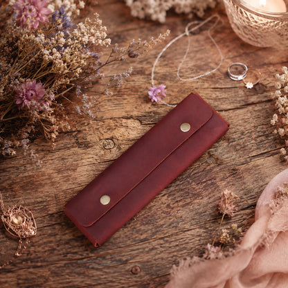 A dark reddish-brown leather ring roll or wallet with two gold-tone snap buttons, styled on a rustic wooden surface with dried flowers, rings, and a candle.