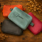 Four leather zippered pouches in orange, mint green, black, and red, arranged on a rustic wooden surface with dried flowers. The mint green pouch in the center features the name 'Genivere' embroidered in black script.