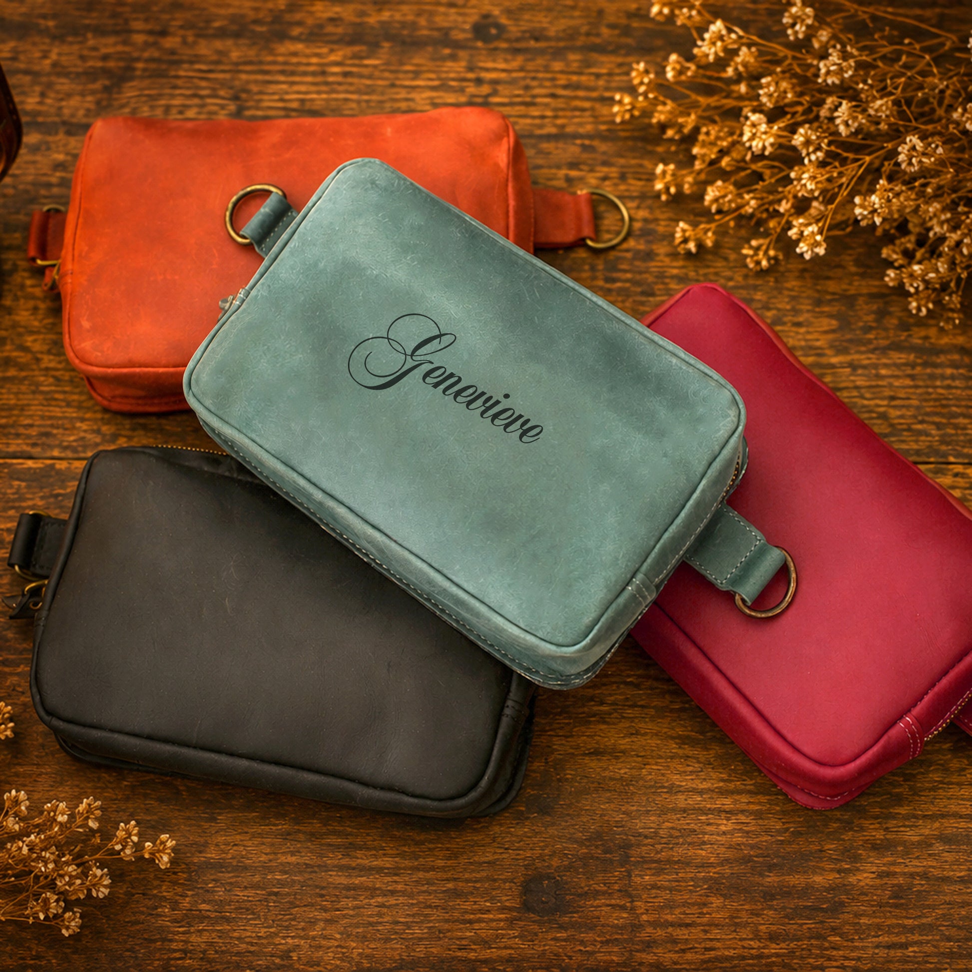 Four leather zippered pouches in orange, mint green, black, and red, arranged on a rustic wooden surface with dried flowers. The mint green pouch in the center features the name 'Genivere' embroidered in black script.