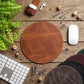 A round brown leather desk mat or mouse pad is centered on a rustic wooden tabletop, surrounded by a keyboard, mouse, earbud case, plant leaves, dried flowers, and other circular pads in different colors. No people or animals are present.