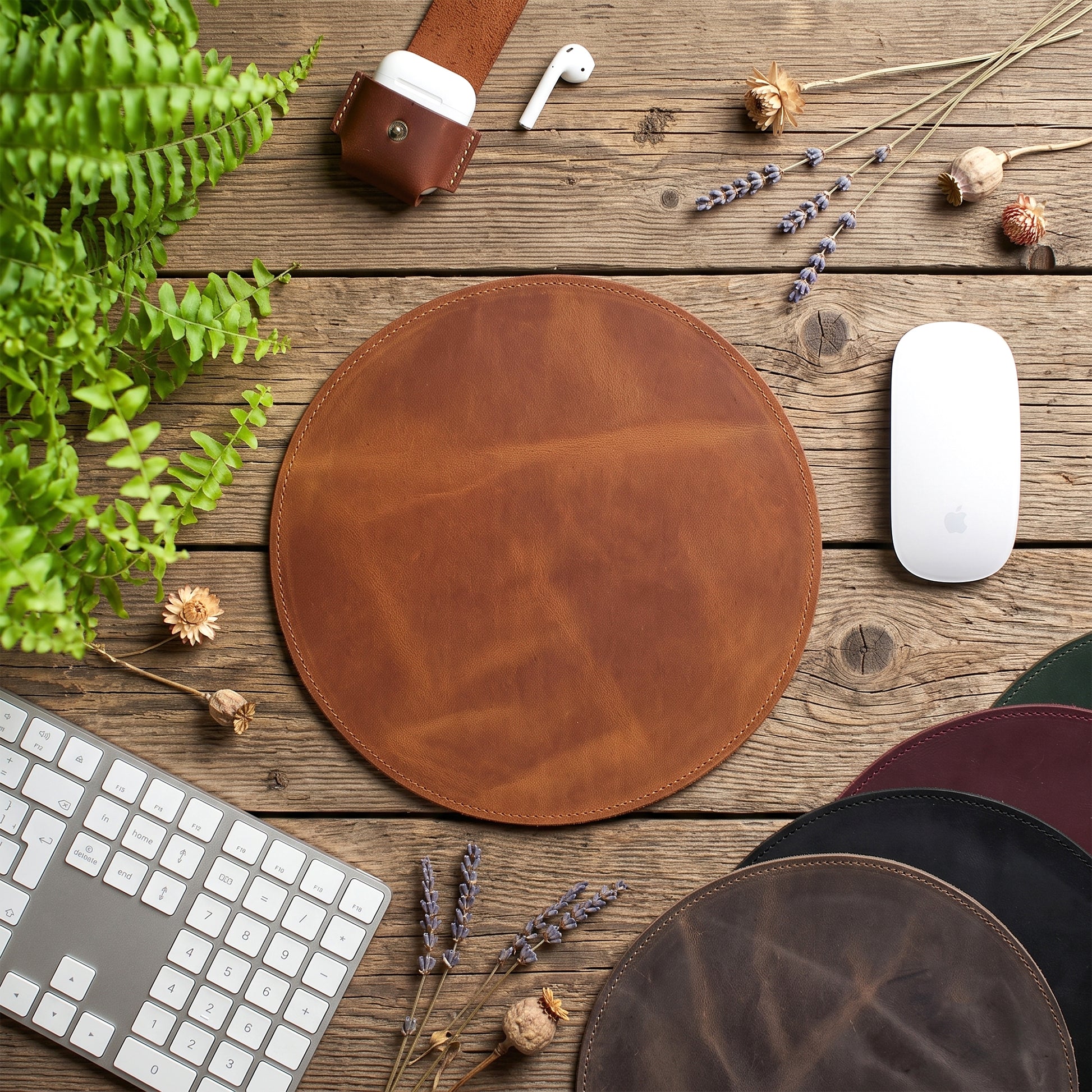 A round brown leather desk mat or mouse pad is centered on a rustic wooden tabletop, surrounded by a keyboard, mouse, earbud case, plant leaves, dried flowers, and other circular pads in different colors. No people or animals are present.