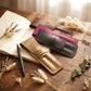 Three personalized leather document pouches in tan, black, and magenta, arranged on a rustic wooden desk with an open notebook, pen, and dried botanicals. The tan pouch is in the foreground and features an embossed monogram logo.