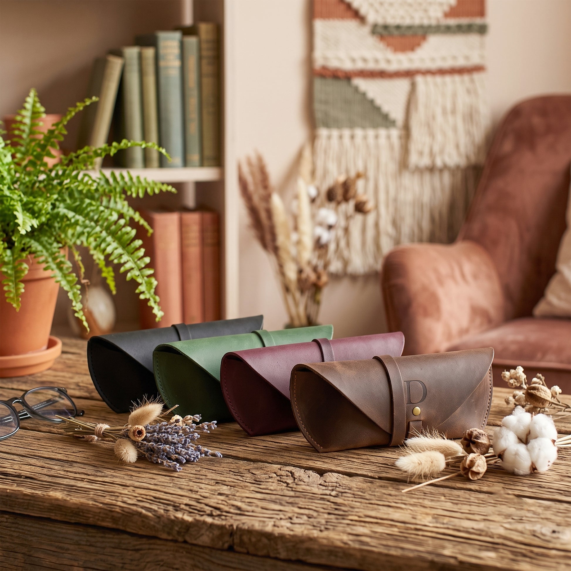 Four leather eyeglasses case sleeves in black, green, burgundy, and brown, arranged on a rustic wooden table in a cozy, boho-style room with a macrame wall hanging, armchair, and potted fern.