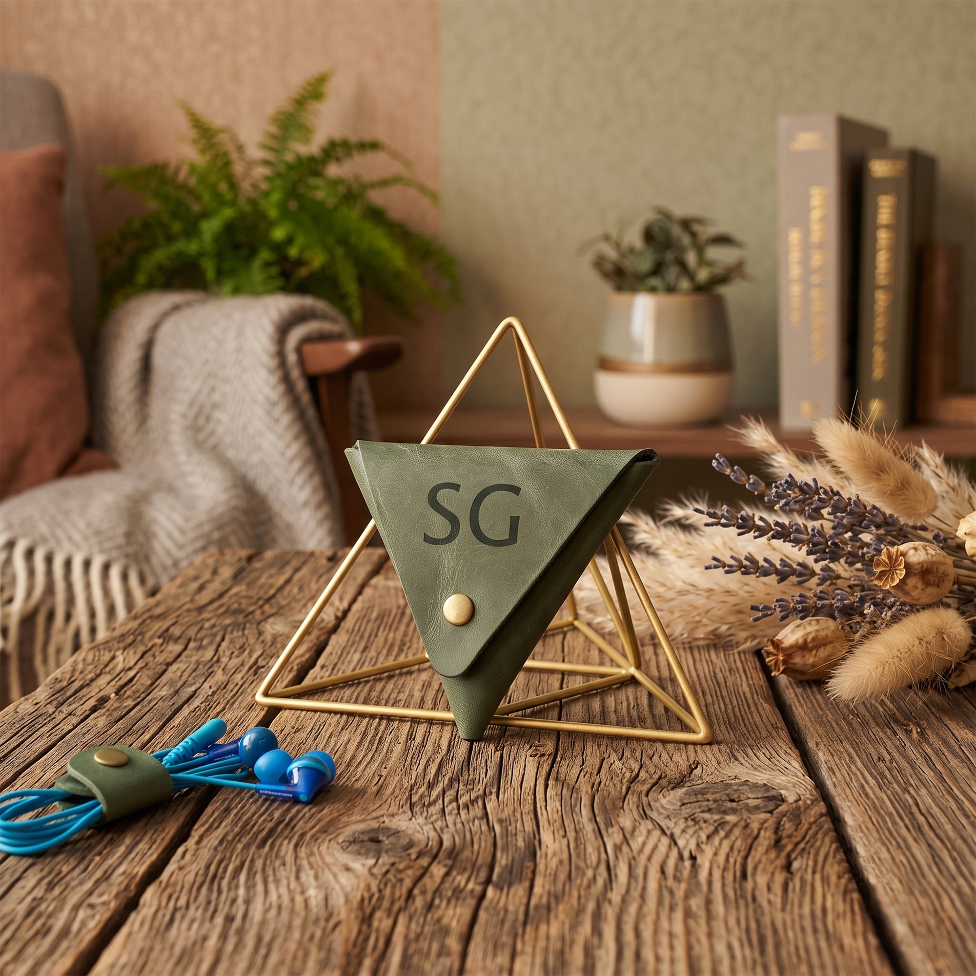 An olive green triangular desk organizer with the initials 'SG' in black, displayed on a rustic wooden table in a cozy home office setting with a gold wire frame stand, blue earbuds, and dried lavender.