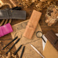 A styled flat-lay of leather roll-up pen cases on a rustic wooden table, surrounded by calligraphy papers, black pens, a pink case, dried flowers, and a glass jar with fairy lights. The central product is a tan leather case with a brass snap and the brand name 'BECK' embossed on it.