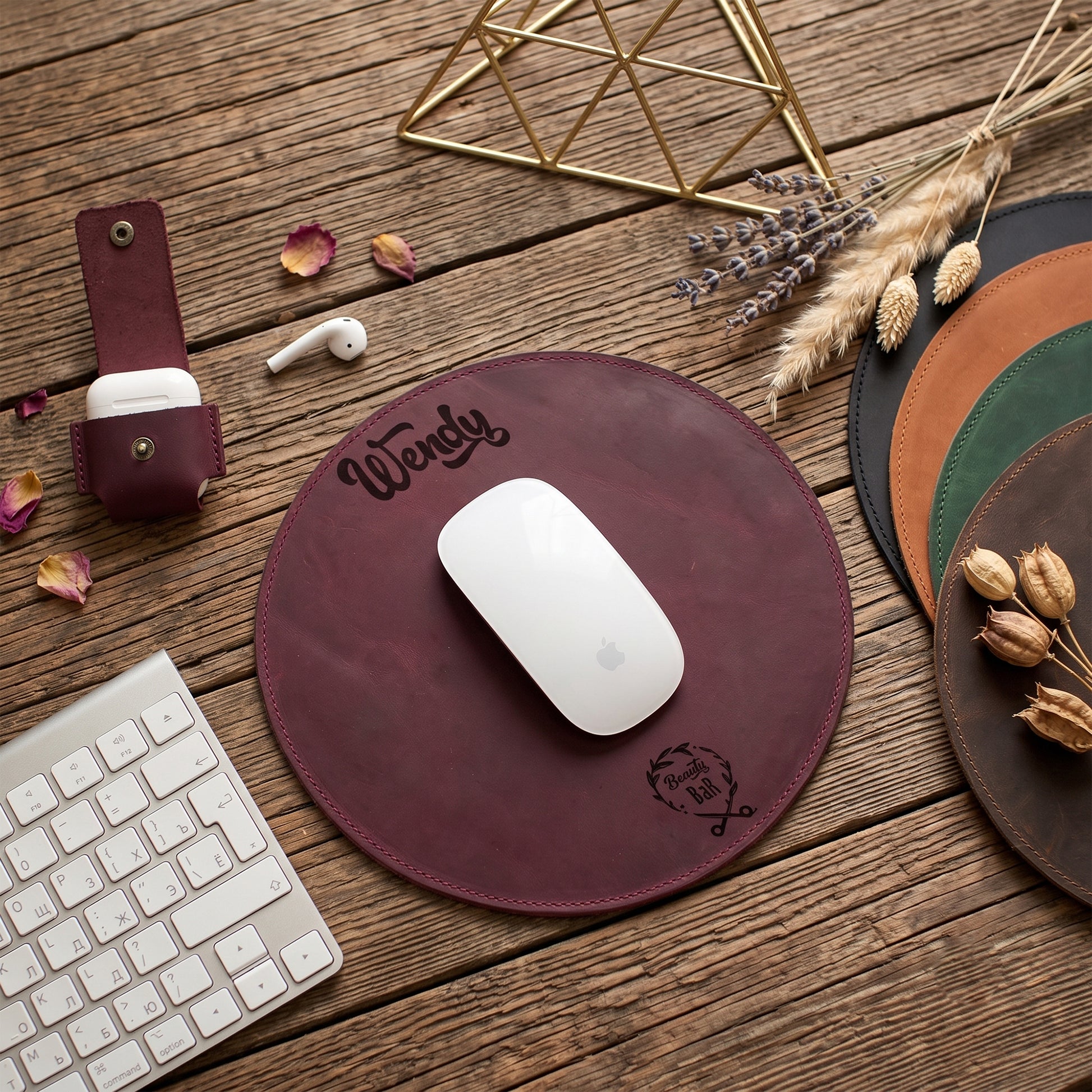 A round burgundy leather mouse pad with 'Wendy' and a monogram logo, styled on a rustic wooden desk with a white mouse, keyboard, earbuds case, and other desk accessories.