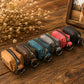 Six small zippered pouches in assorted colors are arranged in a row on a wooden surface. The pouches include tan, brown, teal, gray, pink, and black, each with a metal key ring and zipper.
