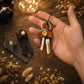 A hand holds a brown leather keychain organizer with keys and a gold snap button, while two additional keychain organizers in dark gray and brown rest on a wooden surface with dried flowers and warm string lights in the background.