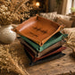 A stack of square leather trays in multiple colors, including tan, green, taupe, burgundy, and black, with the top tan tray engraved with 'I love you' and a soundwave design. The trays are styled on a rustic wooden table with dried flowers and a cozy, warm background.