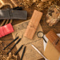A styled flat-lay of leather travel document pouches and accessories on a rustic wooden surface. The central product is a tan leather pouch with a strap and snap closure, embossed with the name 'BECK'.