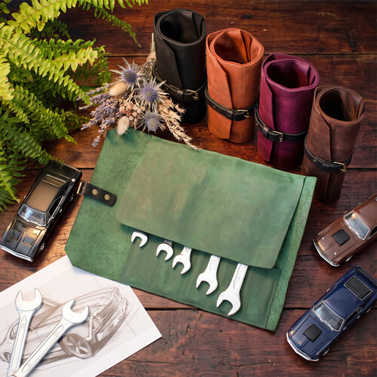 A green leather tool roll with several wrenches partially extended, styled on a wooden tabletop with other rolled tool rolls, toy cars, a fern, and a sketch of a car.