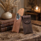 Two leather pouches on a rustic wooden table, one tan and one dark brown, with makeup brushes visible in the tan pouch and the word AGNES printed on the dark brown pouch. Decorative books, a vase of dried flowers, and a softly lit room are in the background.