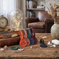 A styled tabletop scene in a cozy living room setting showing several small leather keychain or luggage tags in orange, brown, blue-gray, purple, and black, alongside a black pouch, white earbuds case, and white wireless earbuds. Books, a clock, a glass dome with fairy lights, and dried flowers are arranged around them.